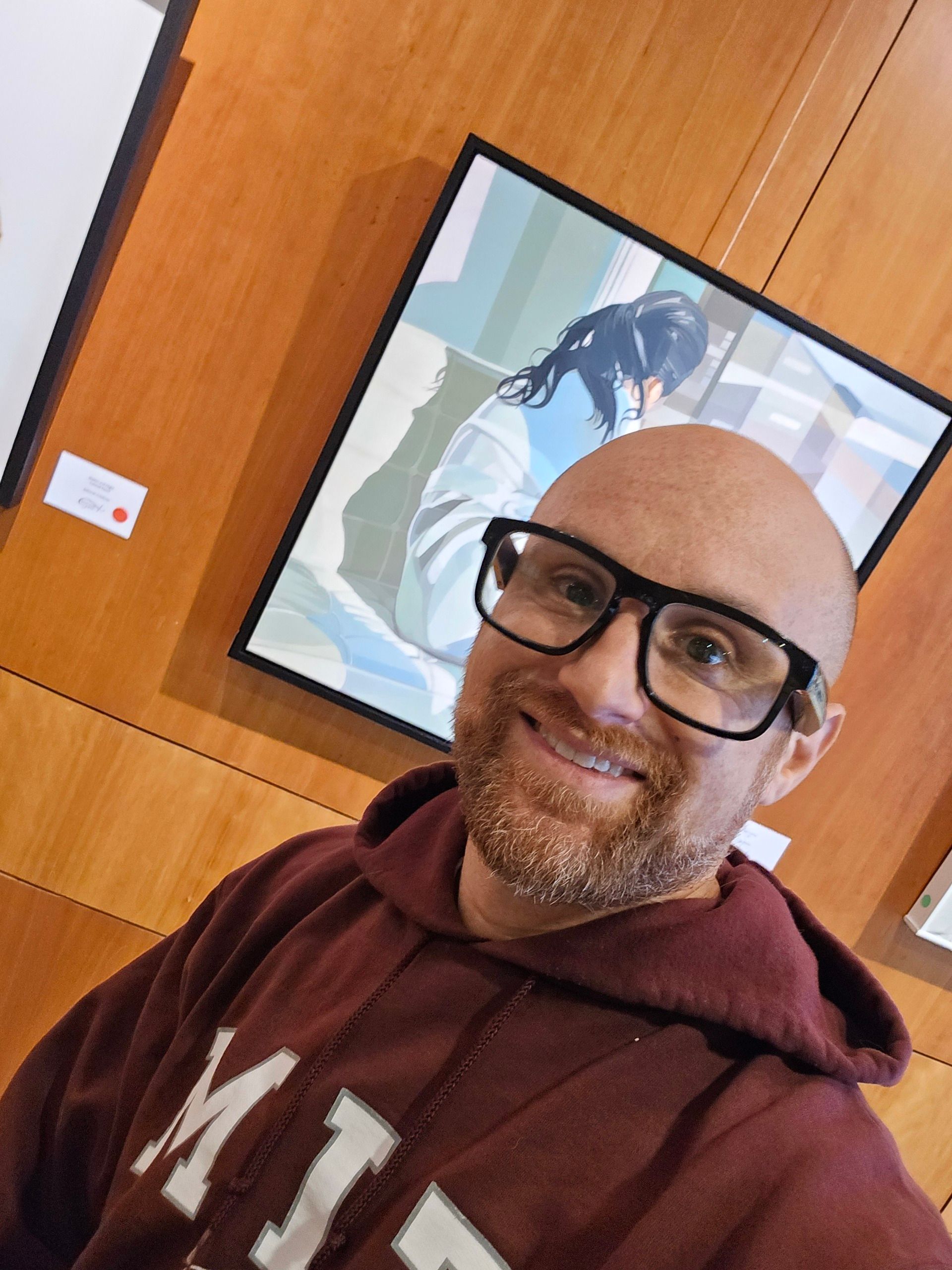 A selfie of me in front of a painting mounted on a wood panel wall. I'm a bald, middle-aged, white man with a red beard flecked with white. I'm wearing glasses with thick black rims and a maroon and white MIT sweatshirt.