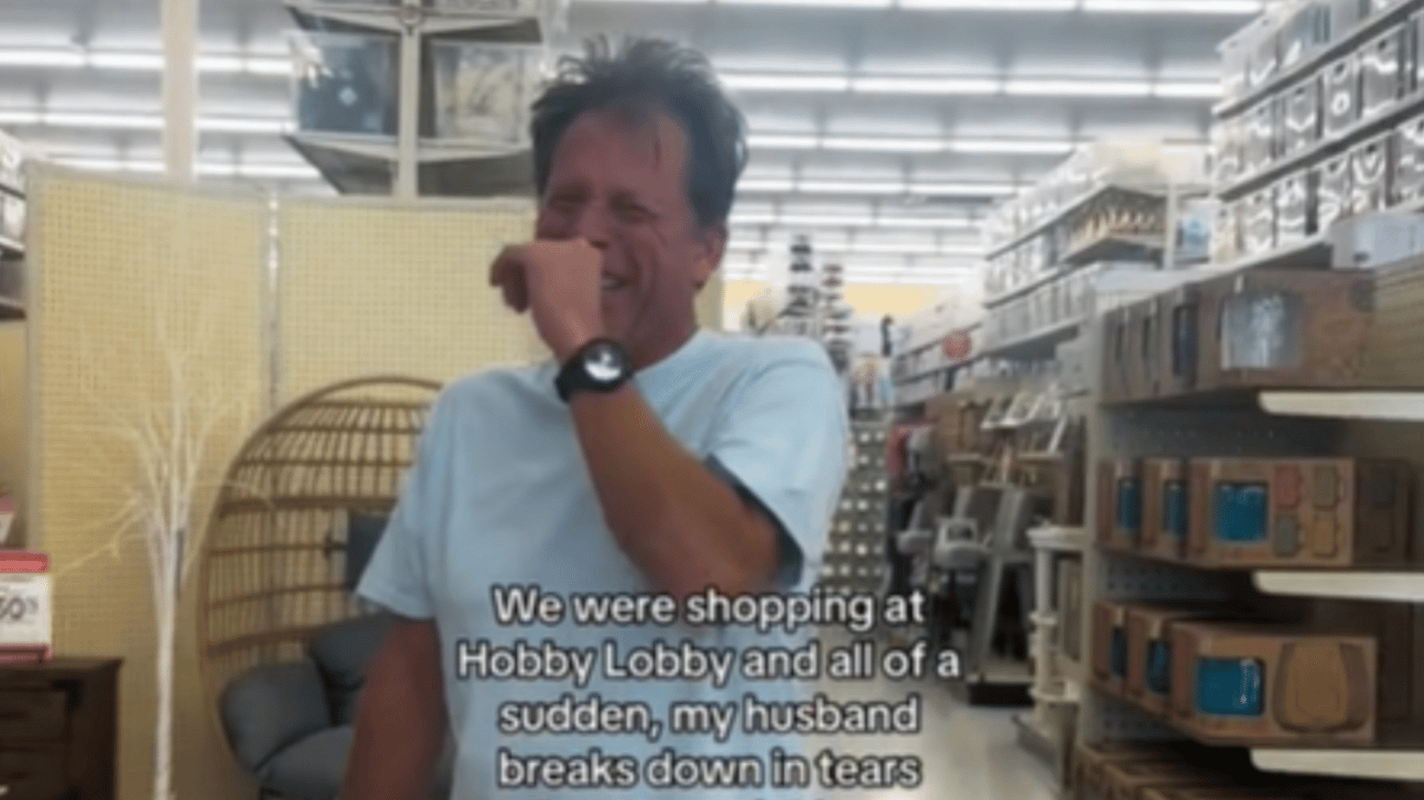 A man stands in a Hobby Lobby, laughing hysterically as he uses a hand to cover his mouth. Text reads: We were shopping at Hobby Lobby and all of a sudden, my husband breaks down in tears
