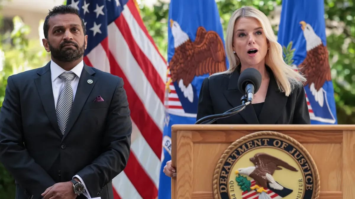 Two officials stand at a podium during a press conference. A woman with long blonde hair, wearing a black blazer, speaks into a microphone at a wooden podium bearing the U.S. Department of Justice seal. A man in a dark suit with a checkered tie and a pocket square stands beside her, looking serious. Behind them are U.S. flags and Department of Justice banners featuring bald eagles.