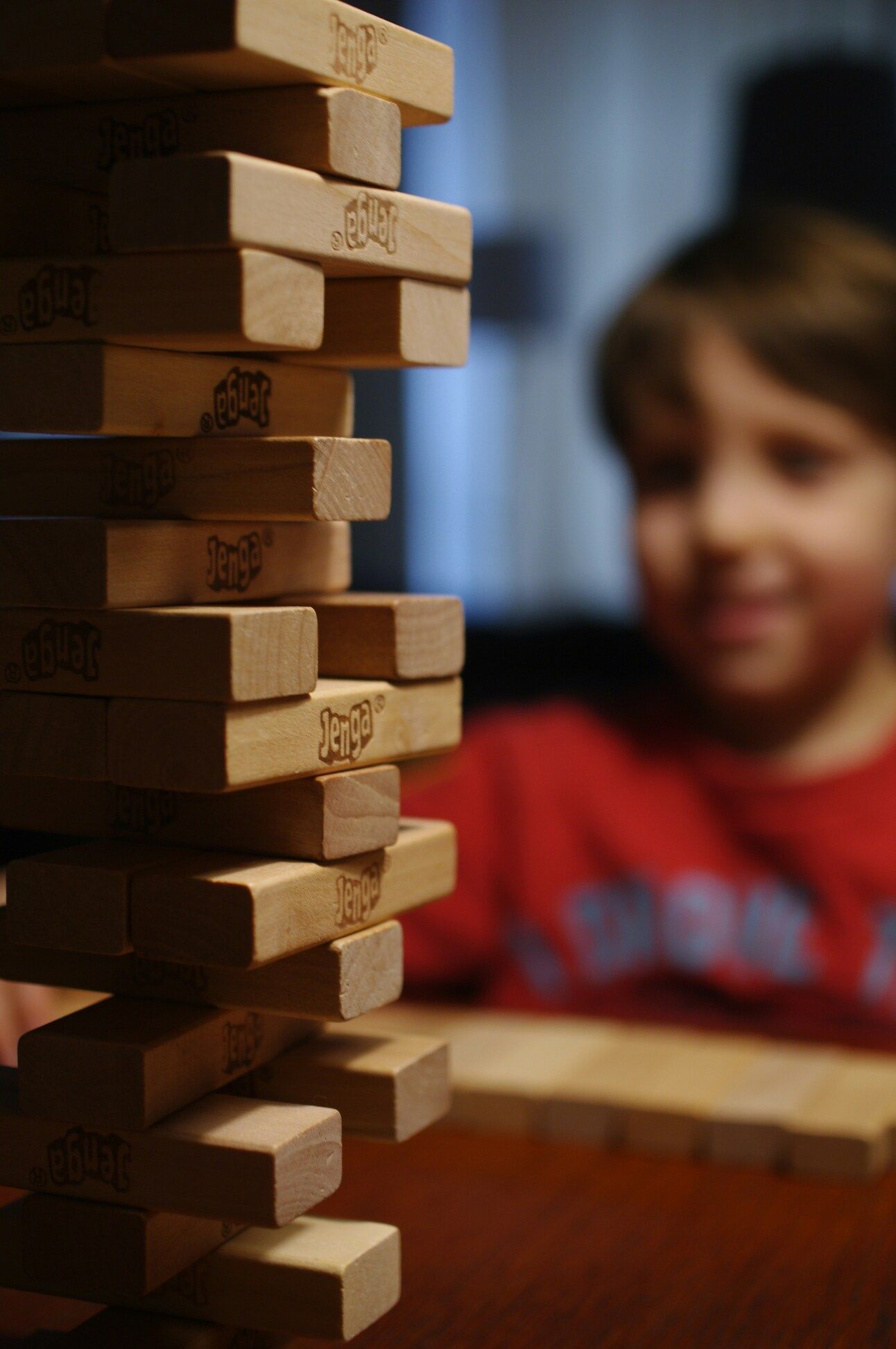 In the foreground is a game of Jenga, with the blocks stacked one on top of the other. In the background out of focus a child is smiling. 