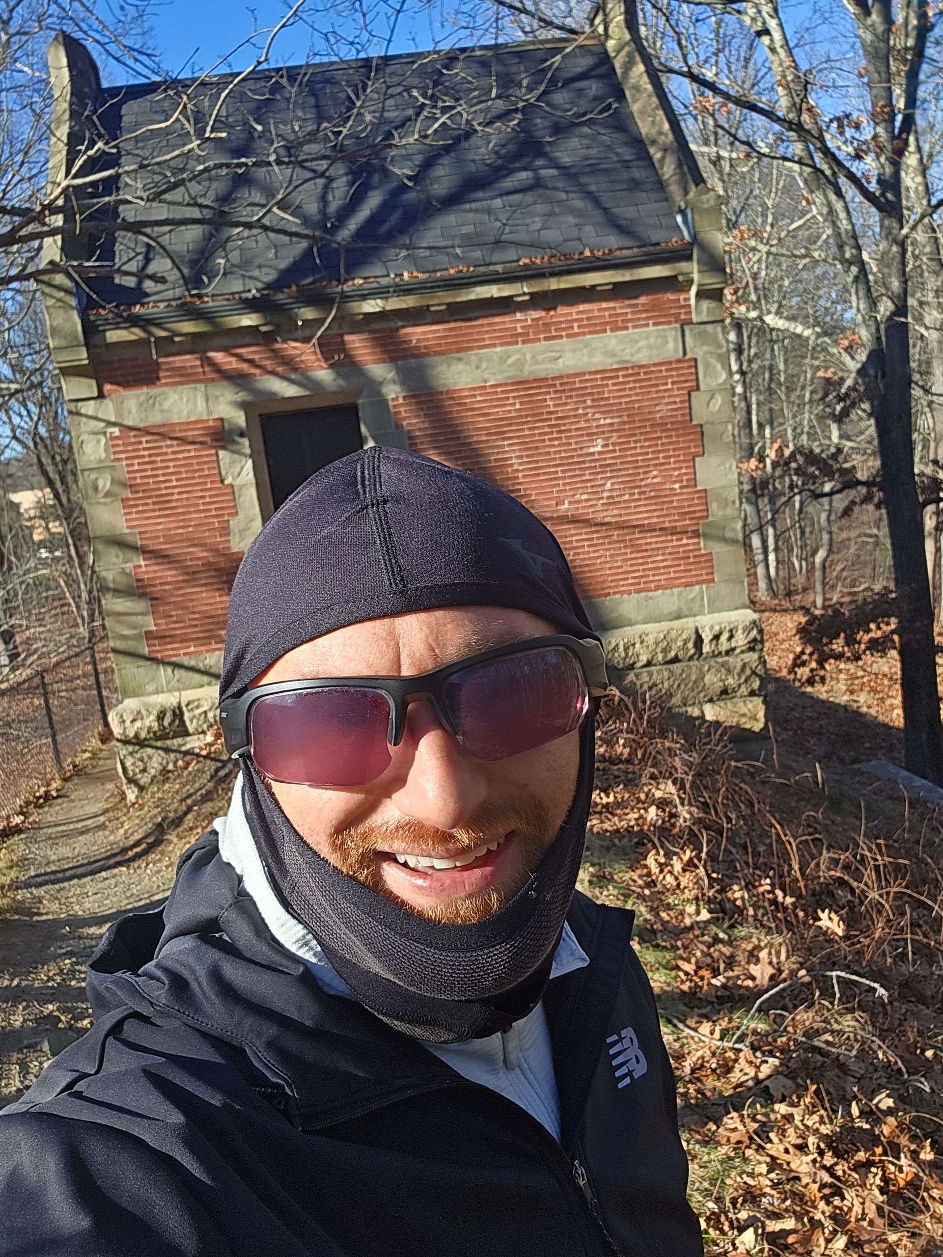 A selfie of me in front of a brick control house on top of an aqueduct on a sunny day. I'm a middle-aged white man with a red beard flecked with white. I'm wearing a black balaclava with the face mask pulled down, fogged up black sunglasses, and a black New Balance running jacked over a light blue running sweater.