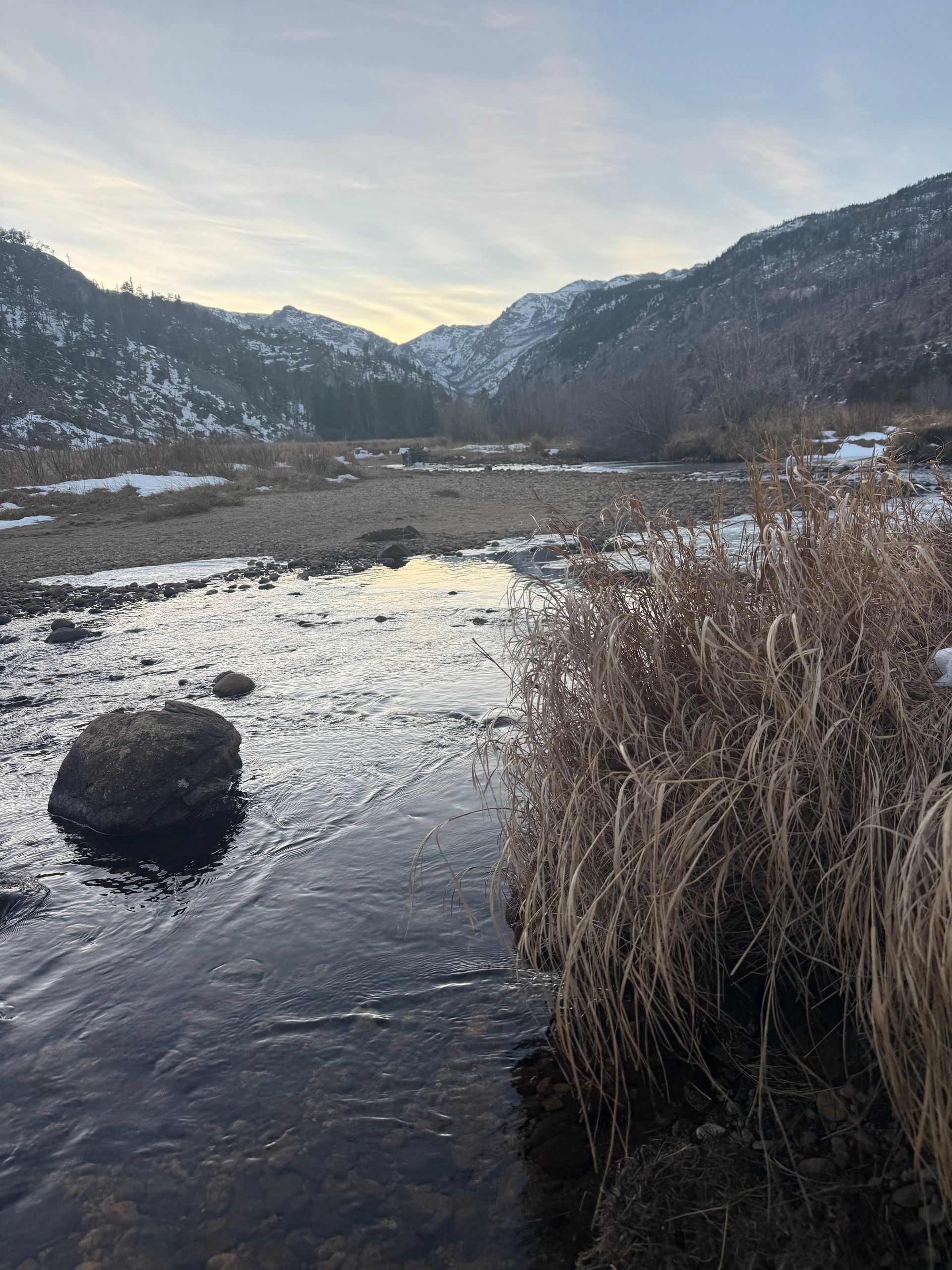 A single boulder sits in a shallow stream flowing through a winter mountain valley, with dried grasses along the bank and snow-capped peaks in the distance under soft morning light.