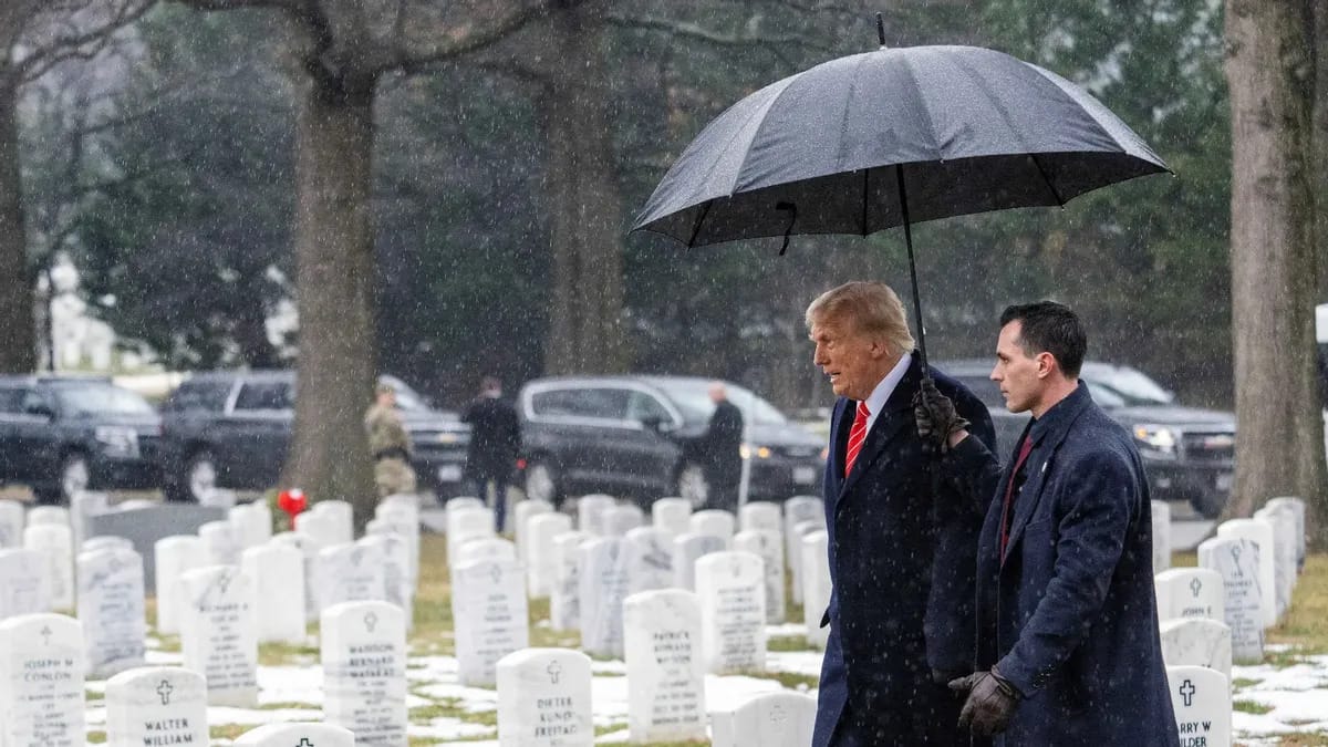 Two white men wearing dark blue suits walk through a cemetary together under an umbrella as it snows.