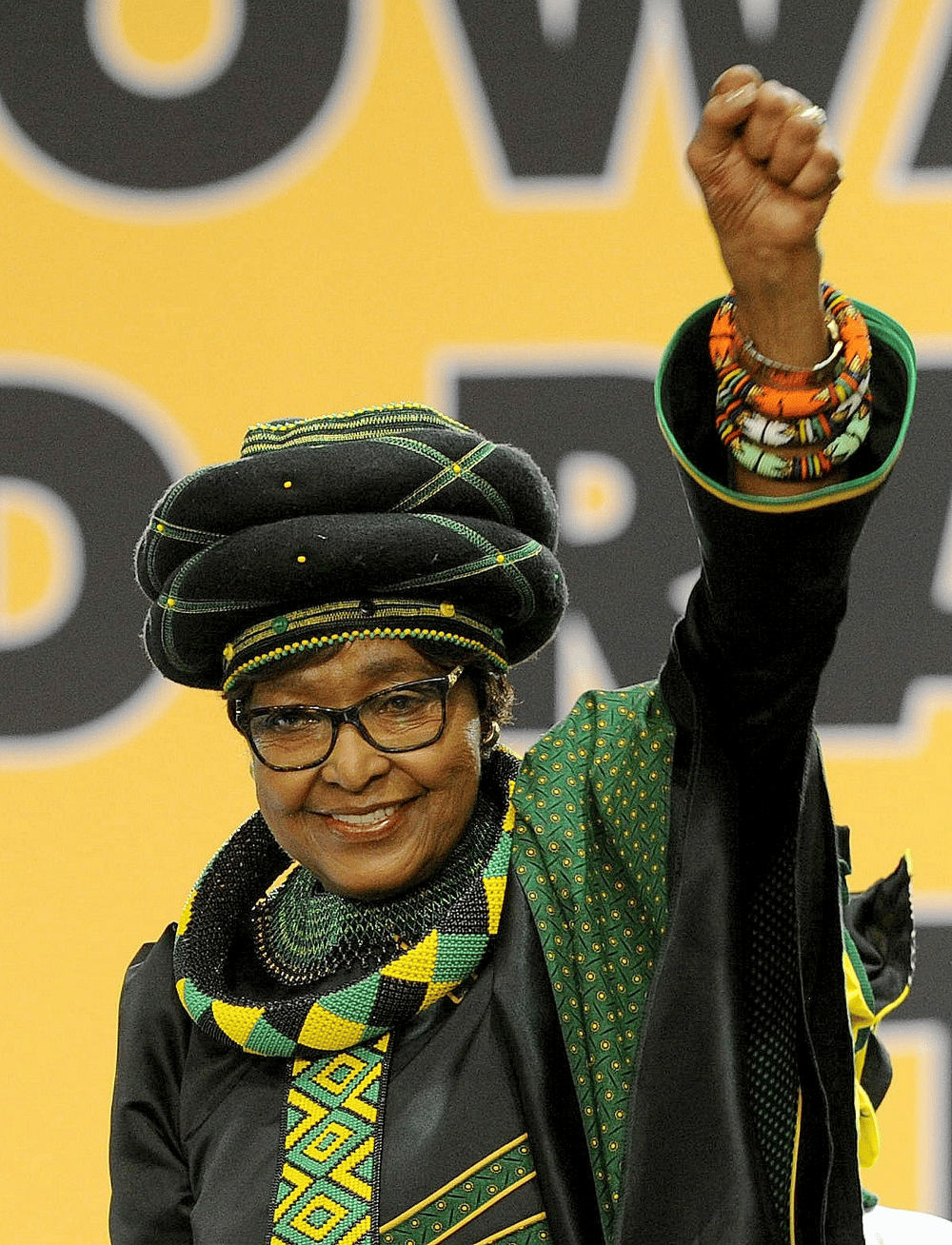 "Winnie Mandela stands in a crowd, her right fist raised in defiance, draped in a black headscarf and patterned shawl. Her face is calm but fierce, lit with the steady flame of someone who has seen too much—and kept going."