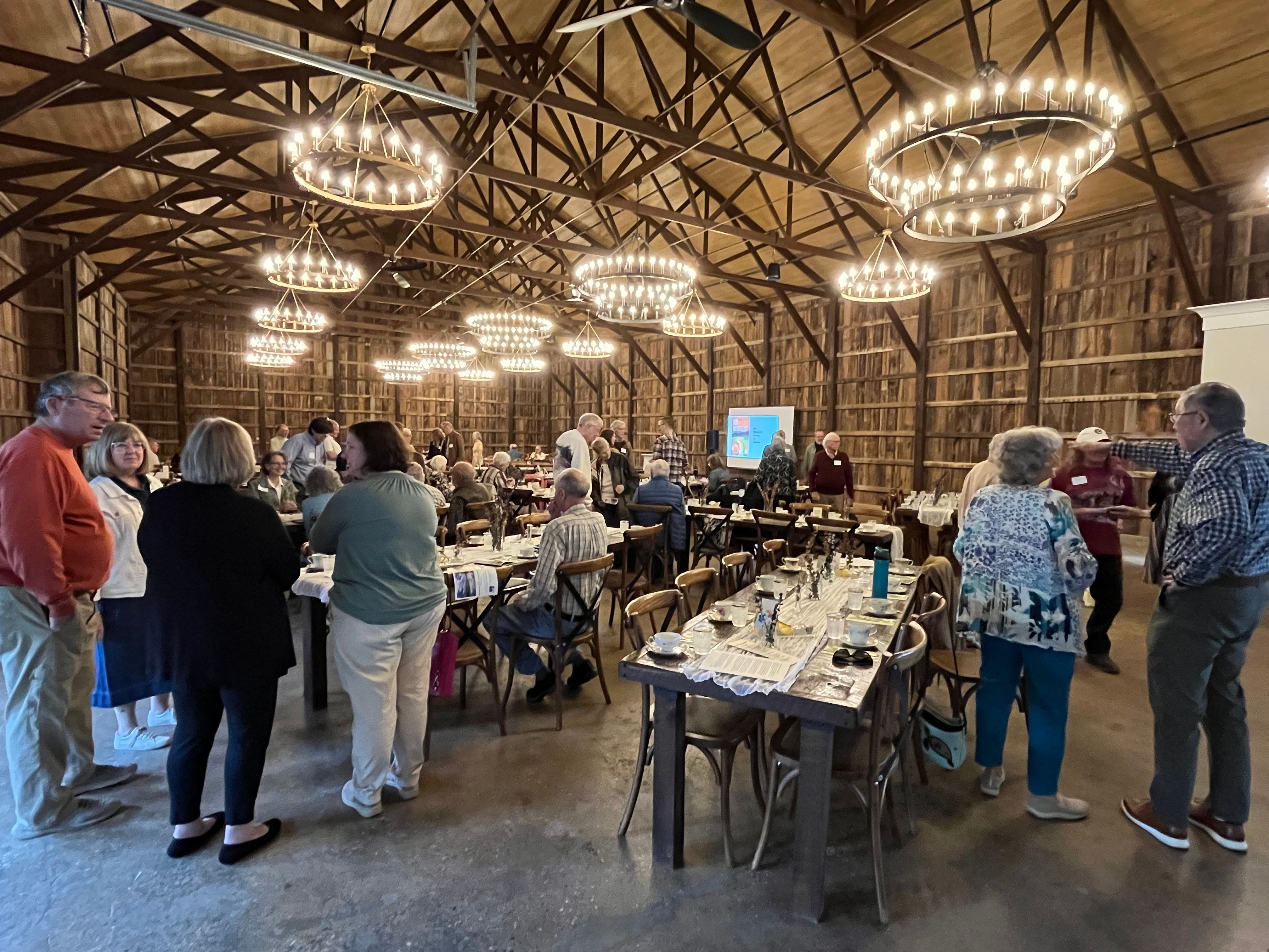 a large converted barn with circular chandeliers and tables of 8 throughout, people milling around, on distant wall a screen for presentation