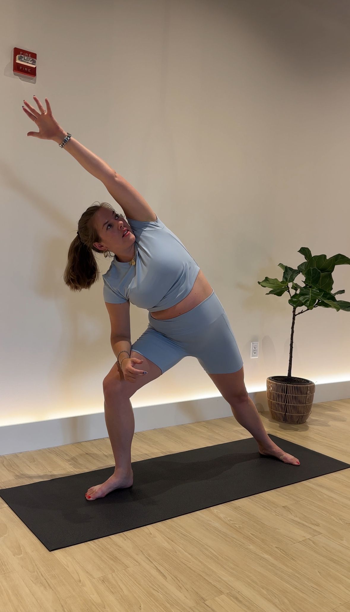 A white woman with a ponytail wears a light blue yoga set with biker shorts and a tshirt. She is in extended side angle yoga pose with one arm long overhead, her front knee bent and her other arm resting on her thigh. She’s in a lightly lit yoga studio on top of a black mat with light oak floors next to a small plant.