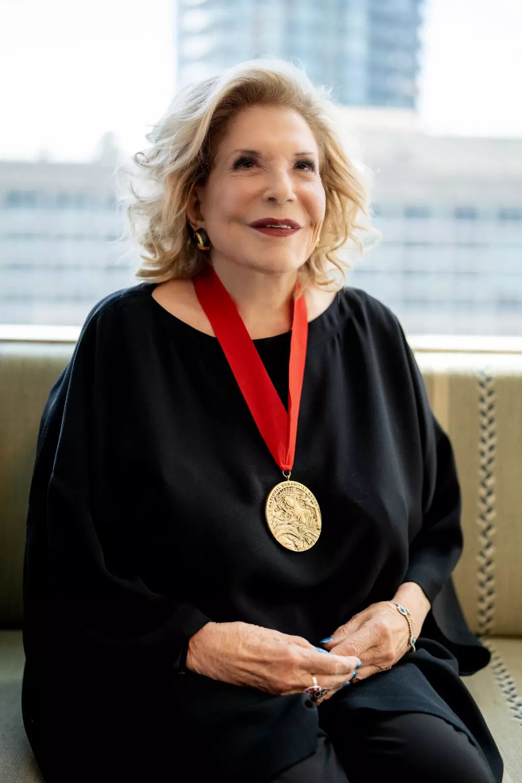 An older woman with wavy blonde hair sits on a couch wearing a black outfit and a large gold medallion on a red ribbon around her neck. She is smiling softly and looking slightly upward. The background shows a cityscape through large windows.