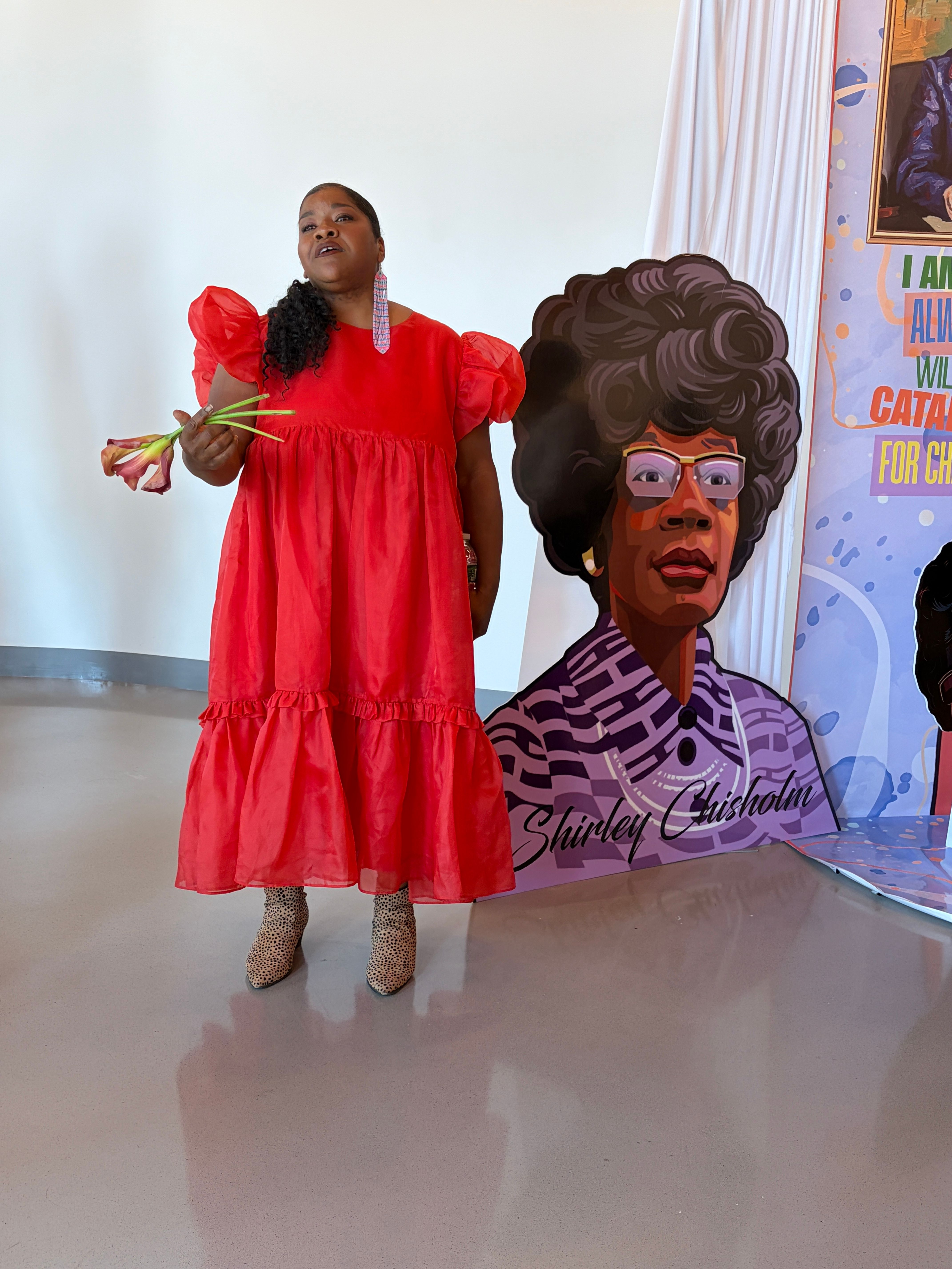 A Black woman in a red dress stands holding pink flowers next to a vibrant portrait of Shirley Chisholm. 