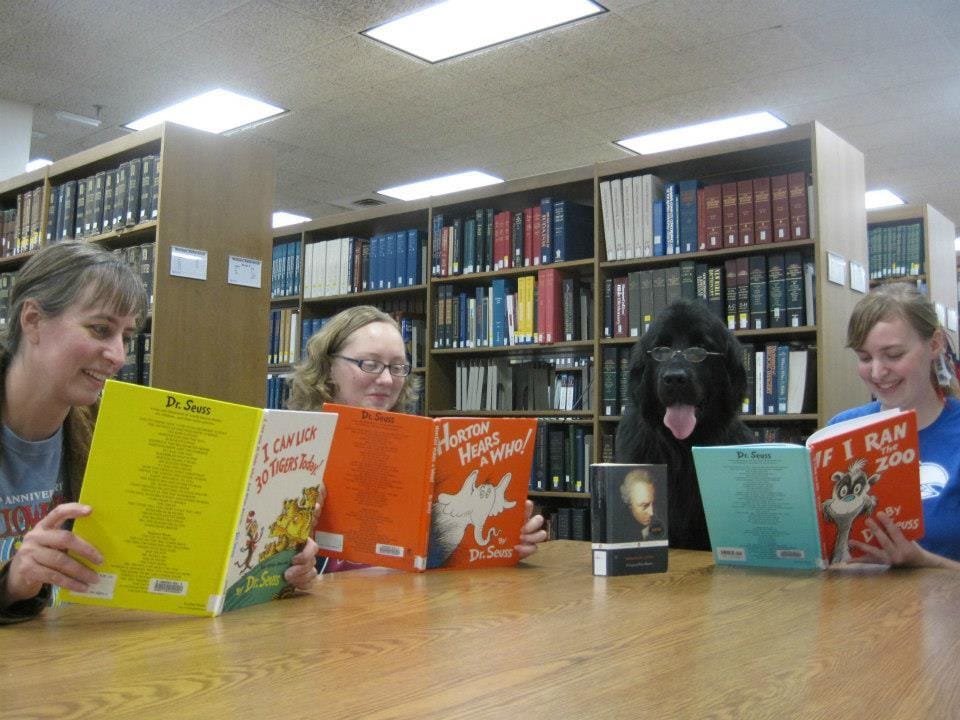 Three white women and one large, black dog wearing glasses sit at a library table. The humans are reading Dr. Suess book while the dog has a philosophy book in front of her. There are bookshelves behind them. It is exactly as silly as it sounds.
