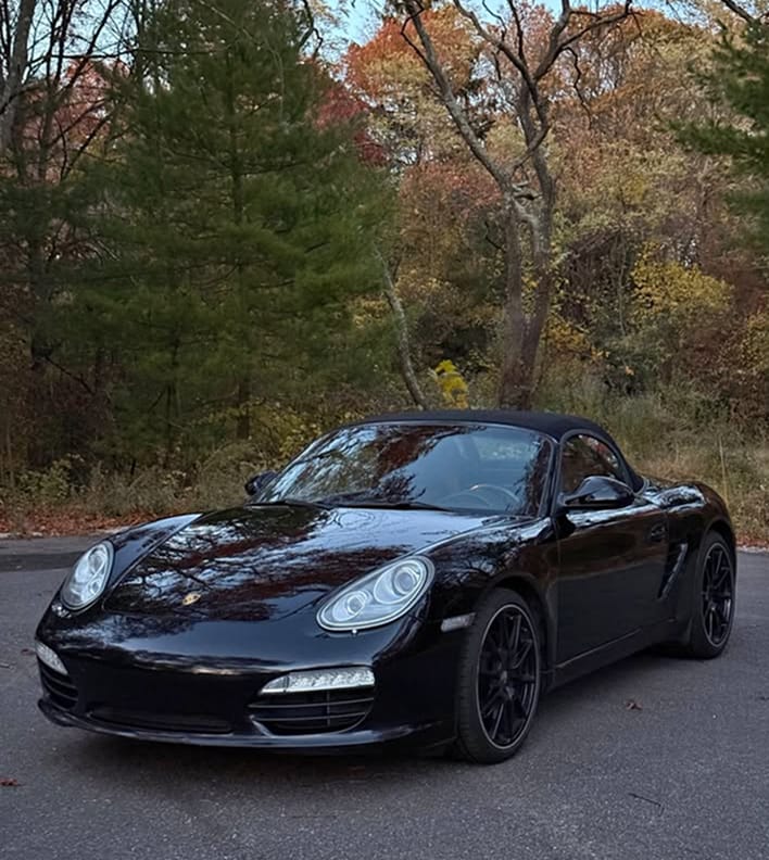 Front three-quarter view of an all-black 2012 Porsche Boxster S with black wheels, parked on a road with autumn trees in the background