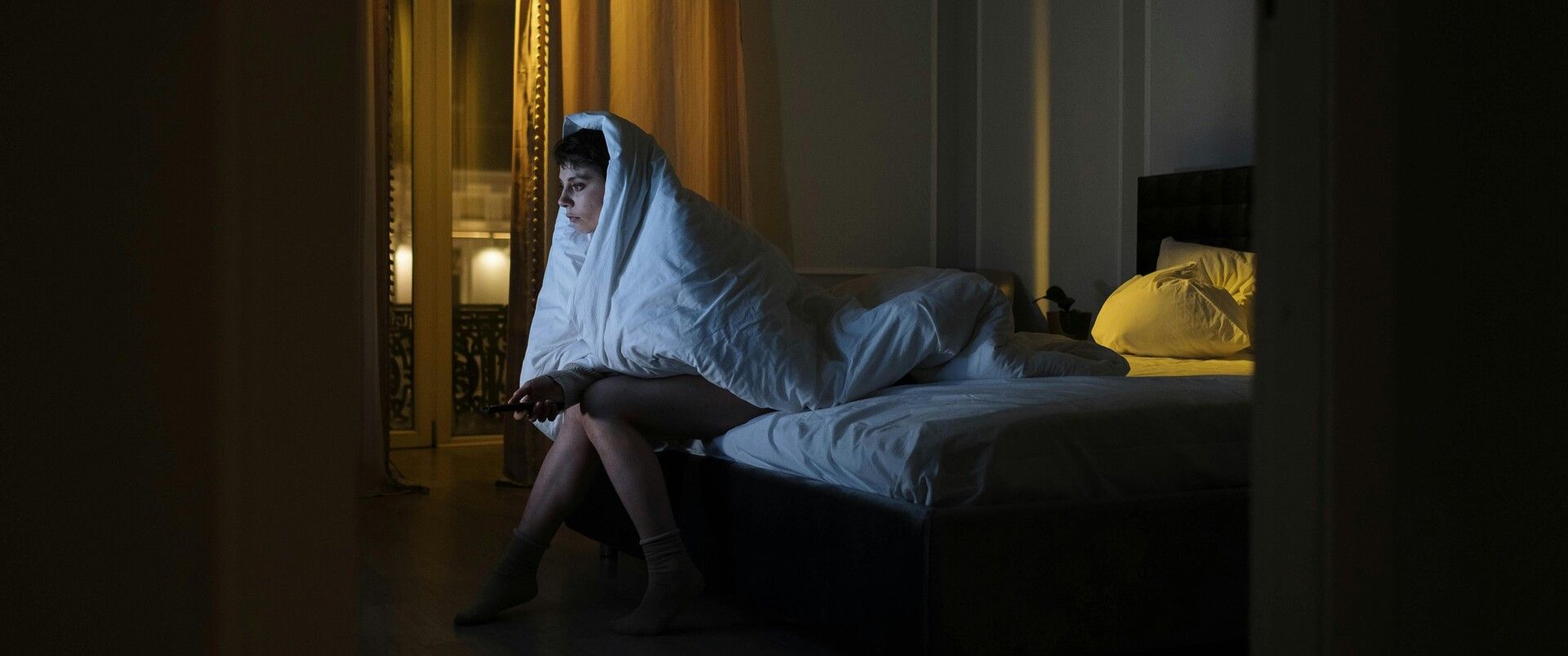 Woman sitting on a bed under a blanket, watching TV as a way to avoid stress and escape problems.