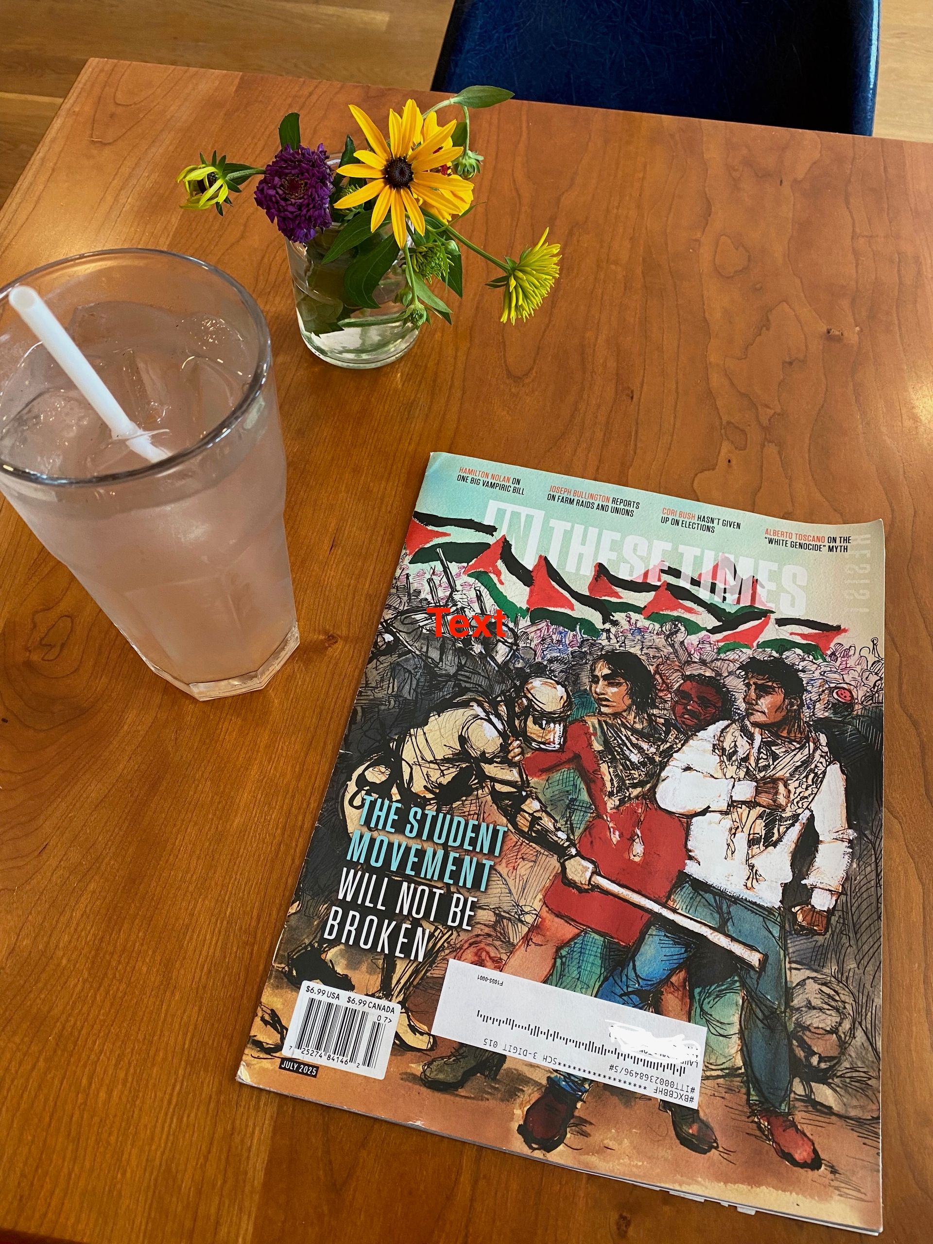 An issue of In These Times on a cafe table next to an iced drink and a jar of flowers.