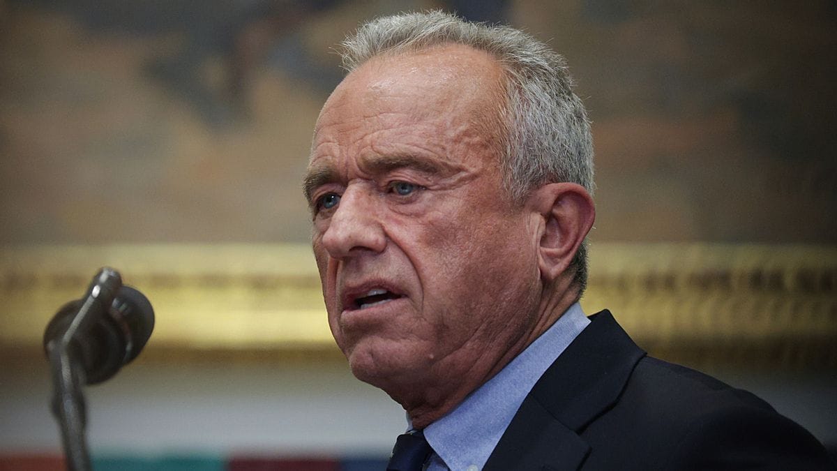 A white man wearing a black suit stands to the right of a microphone.