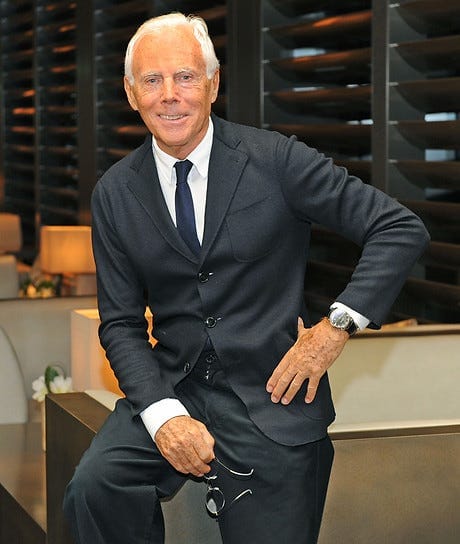 A smiling older man with white hair wearing a dark suit, white shirt, and tie, sitting casually indoors with one hand on his hip and holding eyeglasses in the other.