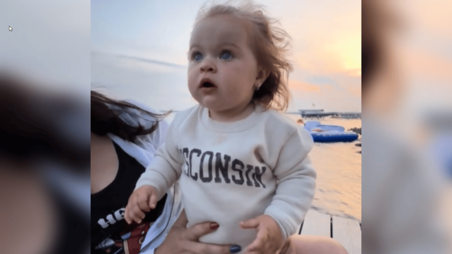 A little girl, eyes wide, looks up into the distance at something we can't see. She's out near some water and is being held by an adult