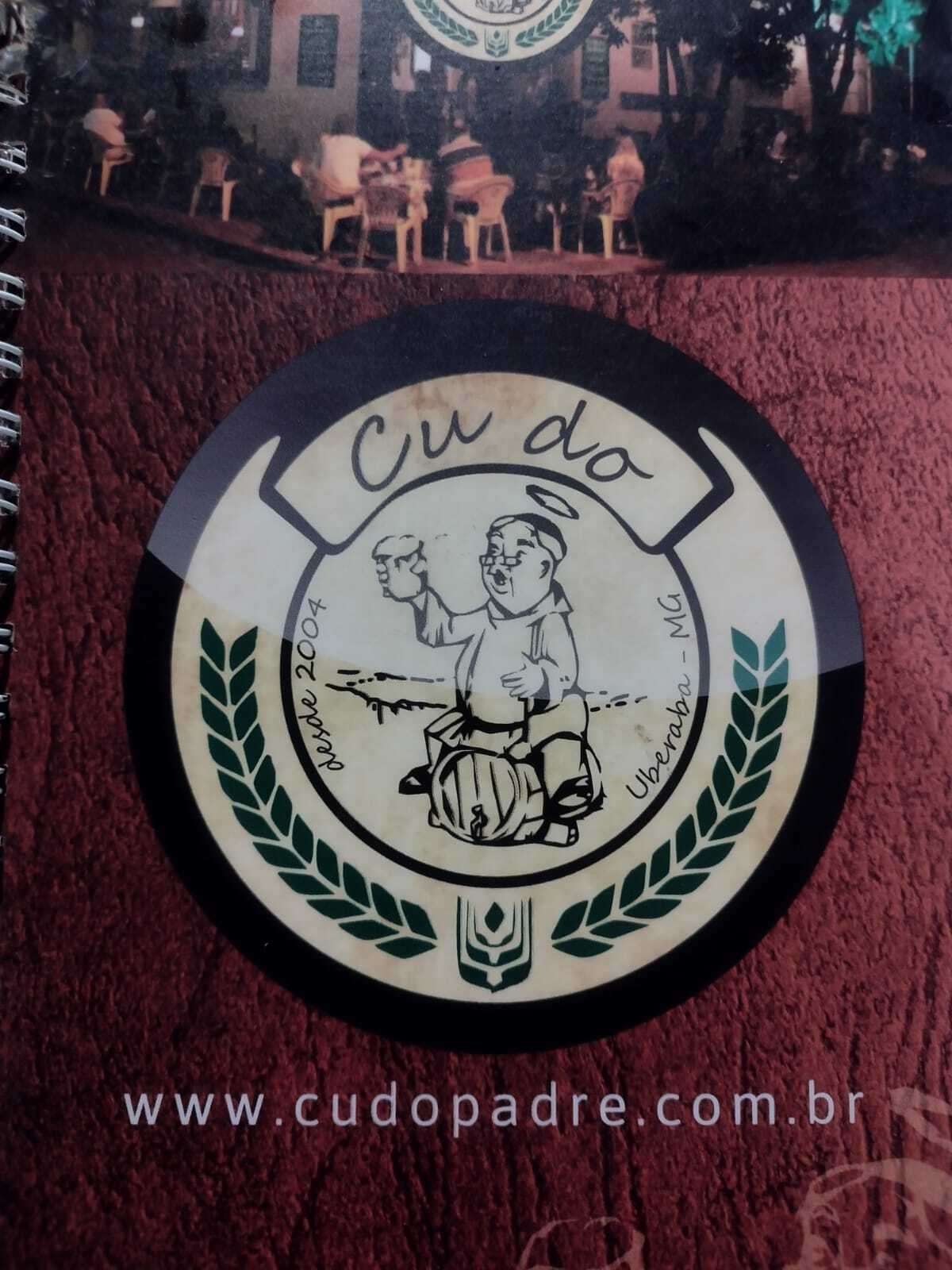 Foto do cardápio da Chopperia Cu do Padre, em que tem um círculo, dois ramos de cevada, um padre sentado cm um copo de serveja, e a inscrição Cu do Padre