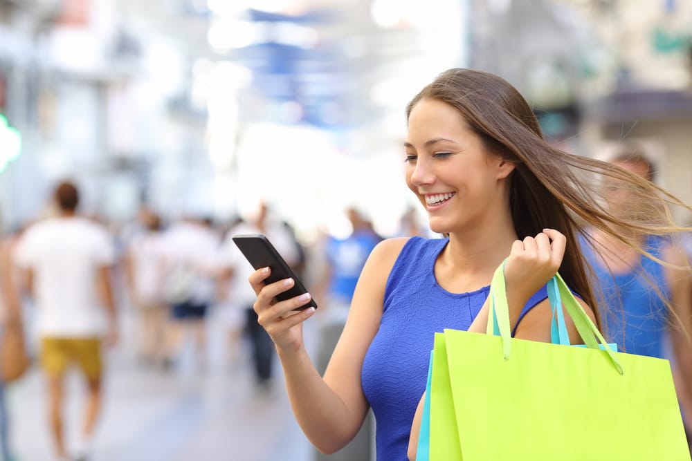 woman holding shopping bags looking at smart phone walking down busy street vervology 2