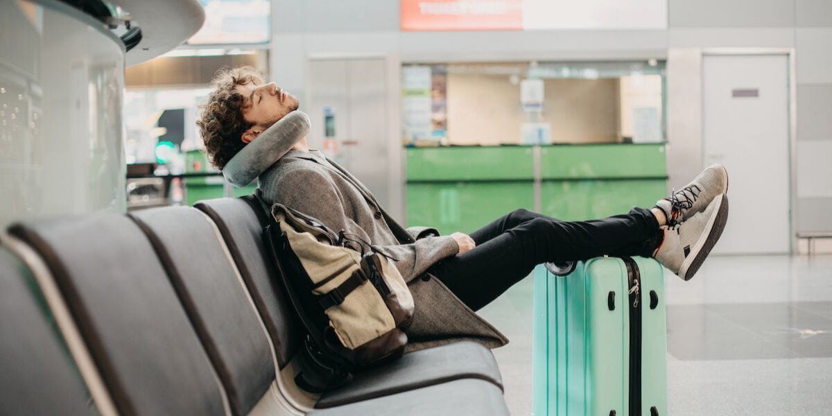 a guy sleeping in an airport with a neck pillow and feet up