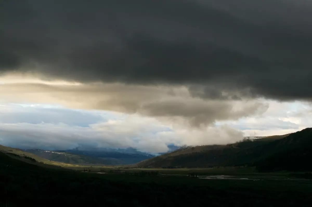 A thick layer of storm clouds gathers with blue sky and hills visible in the background, with a dark valley in the foreground.