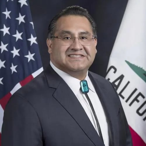 Portrait of Assemblymember James C. Ramos standing in front of the U.S. and California flags, wearing a dark suit, glasses, and a turquoise bolo tie.