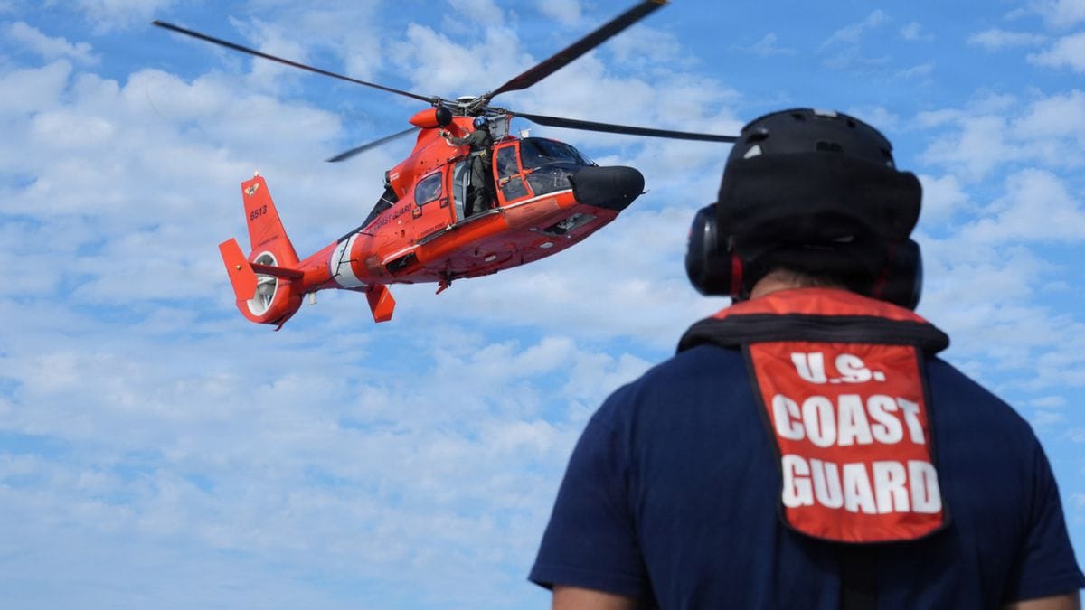 A person looks at a helicopter flying above them. THeir back says U.S> COAST GUARD.