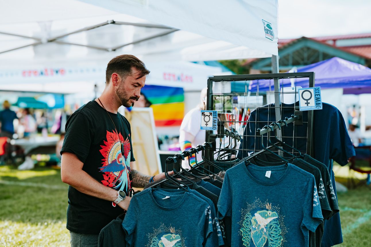 A man earing an Earth Medicine Apparel Co. shirt looks at display racks of shirts. Pride flag in background.
