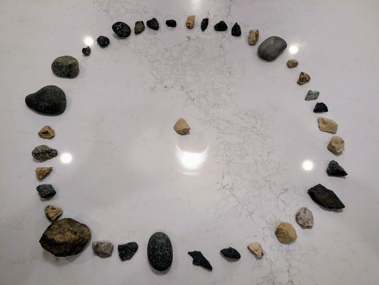 "Thirty-seven assorted rocks arranged in a loose circle on a white marble surface, varying in size, shape, and color, gathered in previous Catharsis Theater sessions."