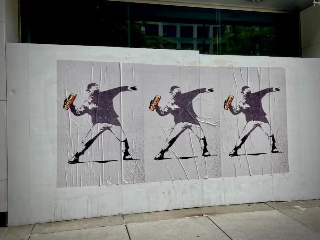 Black image on a white background shows a masked man with a backwards baseball cap about to throw something. In his hands, instead of flowers like the original Banksy artwork, there is a sub sandwich depicted in color. Three posters appear in the photograph.