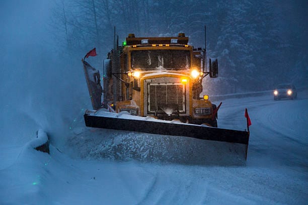 A municipal snow clearing truck plowing snow on a highway early morning with headlights and blade pushing snow off the road