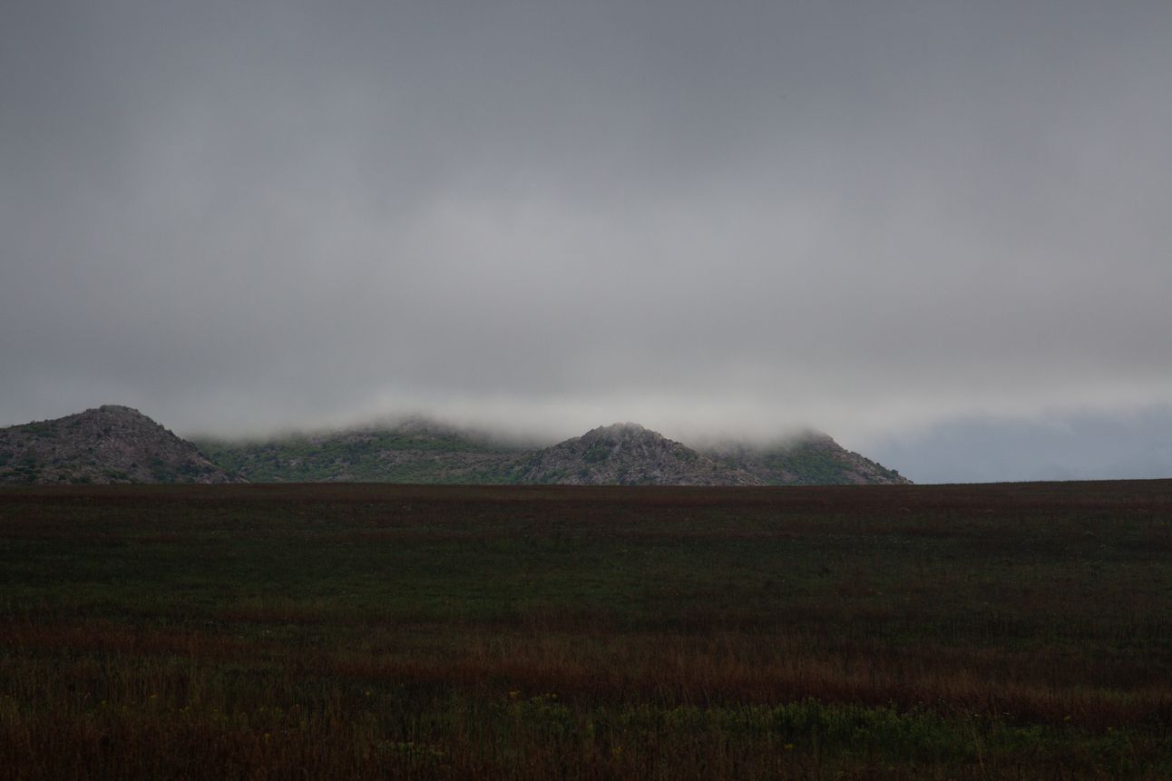 A small hill pokes up in front of a low-hanging grey cloud across a grassy field.