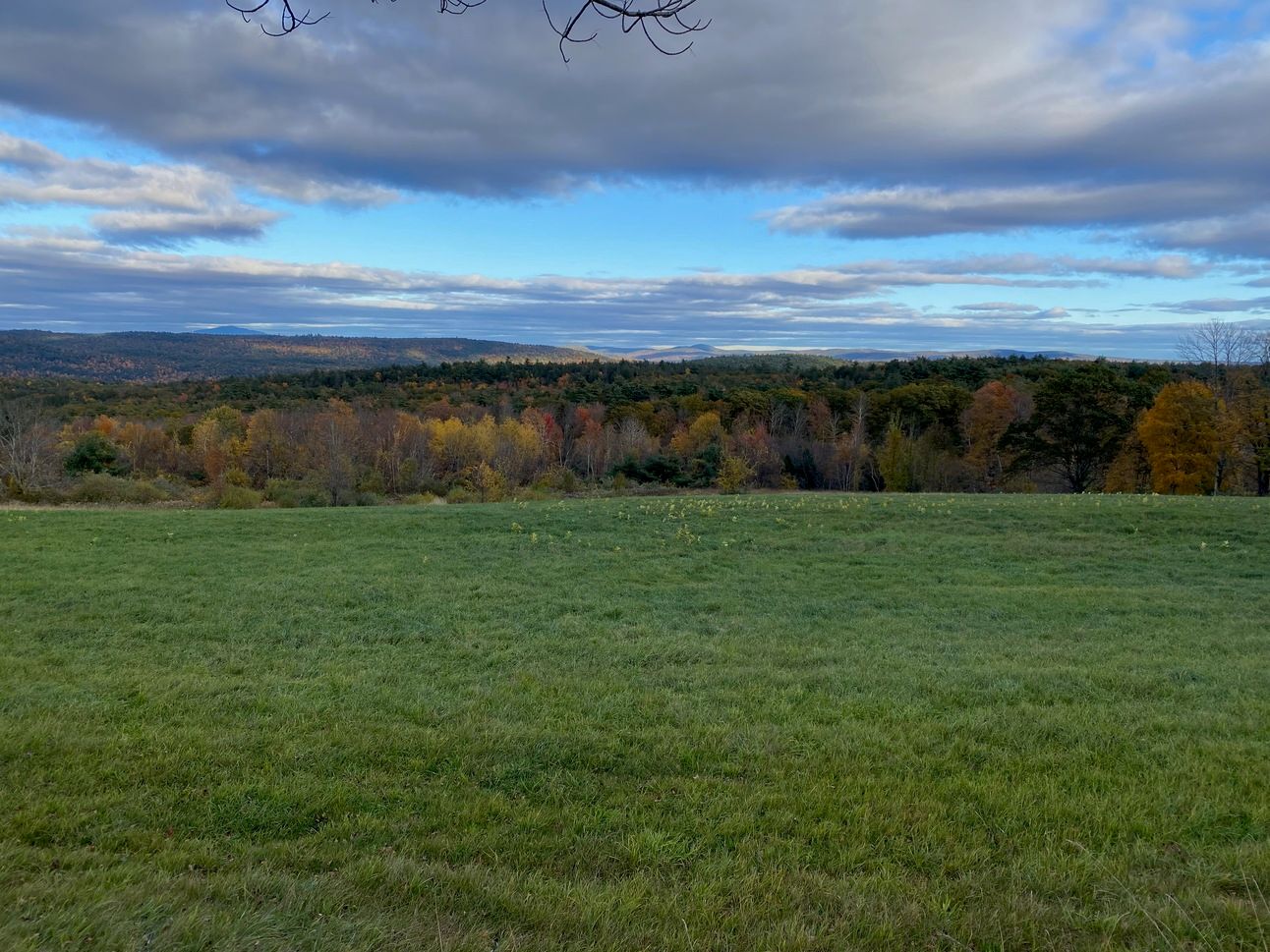 A bright green field with a sweeping view of hills in their fall glory and a sky of silver, blue, and purple clouds.
