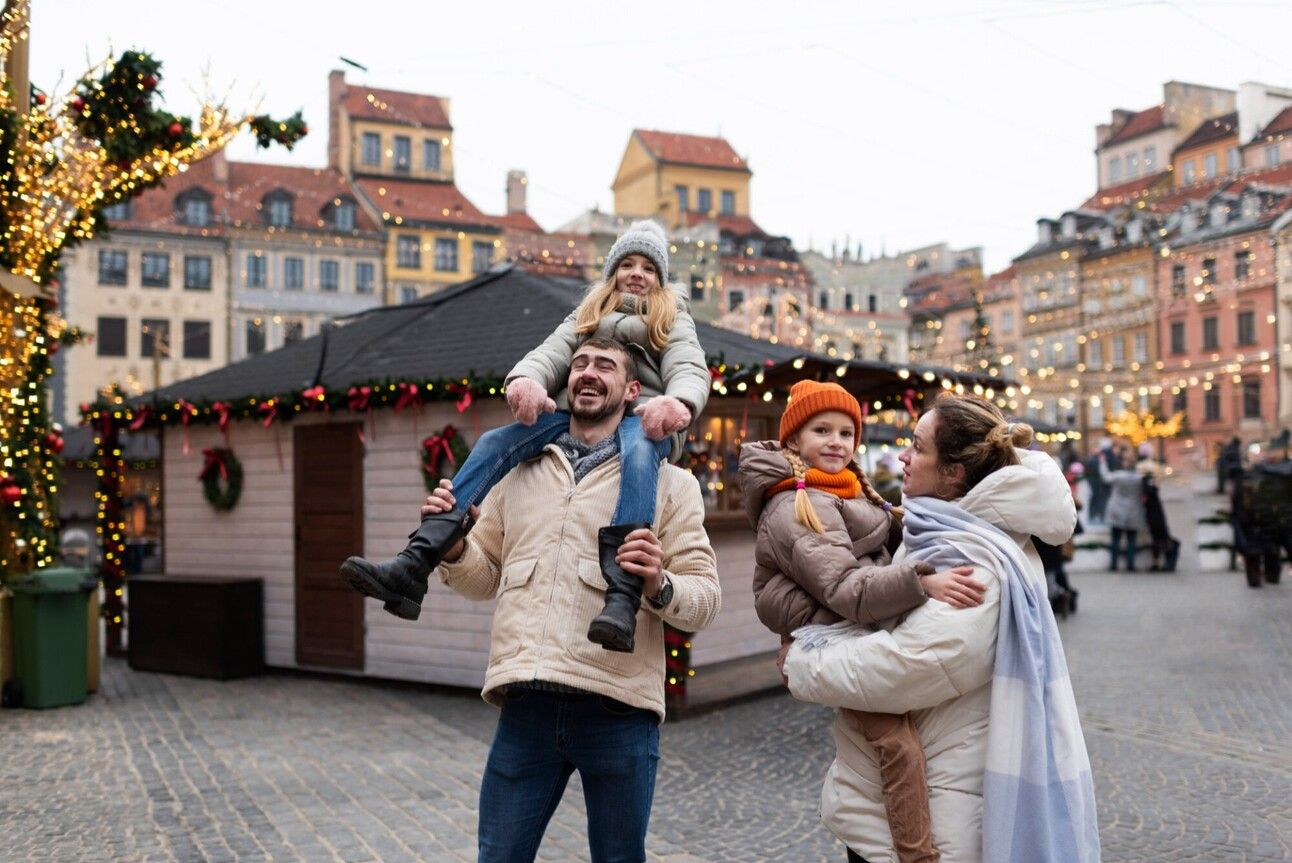 The background is a holiday market with christmas lights and wreaths decorating the booths. In the foreground of the image is a family of a man, a woman and two girls all in winter clothes smiling. The man holds one little girl on his shoulders and the woman holds the other little girl in her arms. 