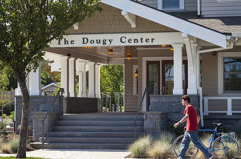 The front entrance of The Dougy Center, a grief support organization, with a young man walking a bicycle past the steps under the center’s white-pillared porch.