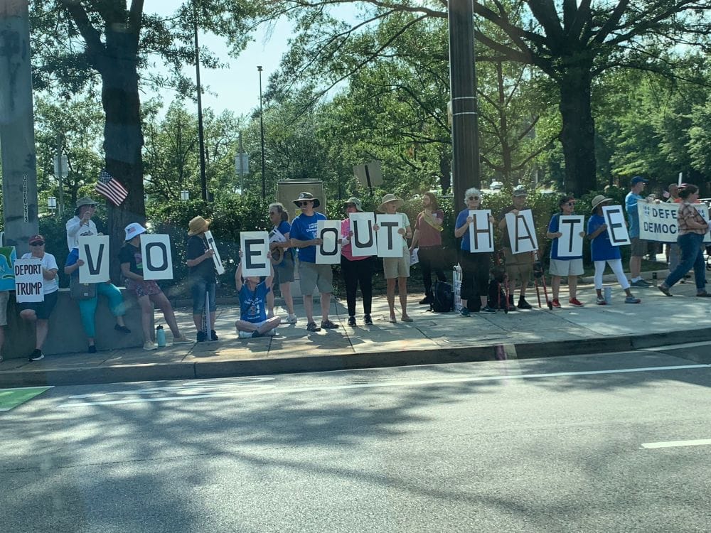 A row of people holding signs on a suburban street corner. Each person holds one big letter, spelling “VOTE OUT HATE.” Trees are visible behind them.