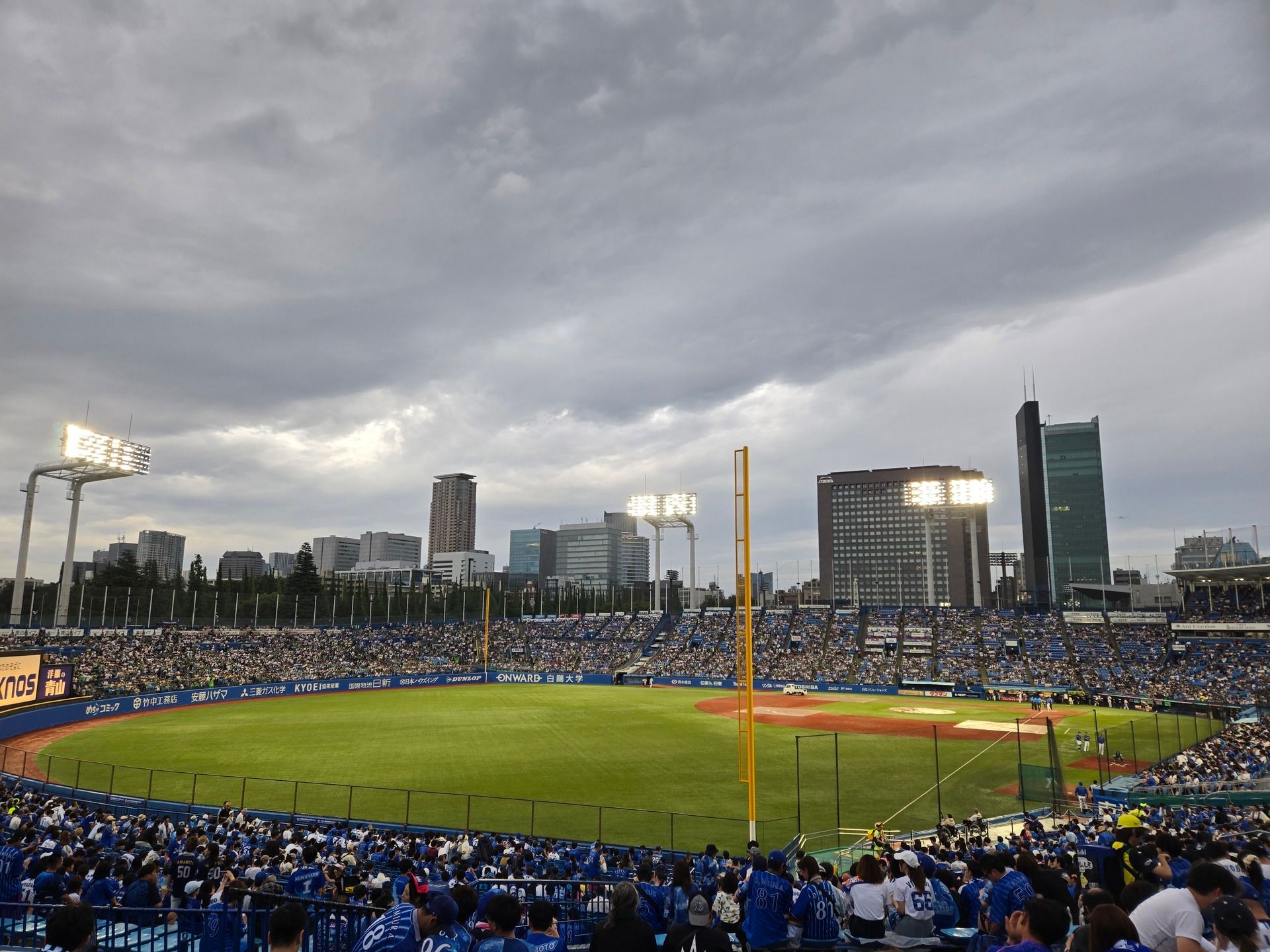 A baseball field in the late afternoon on a cloudy day. A sea of blue shirts are in the near stands, with the skyscapers of Tokyo looming beyond.