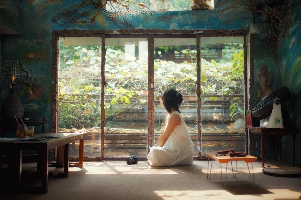 Person sitting cross-legged on the floor in a softly lit artistic room, gazing out a large window covered with green vines.