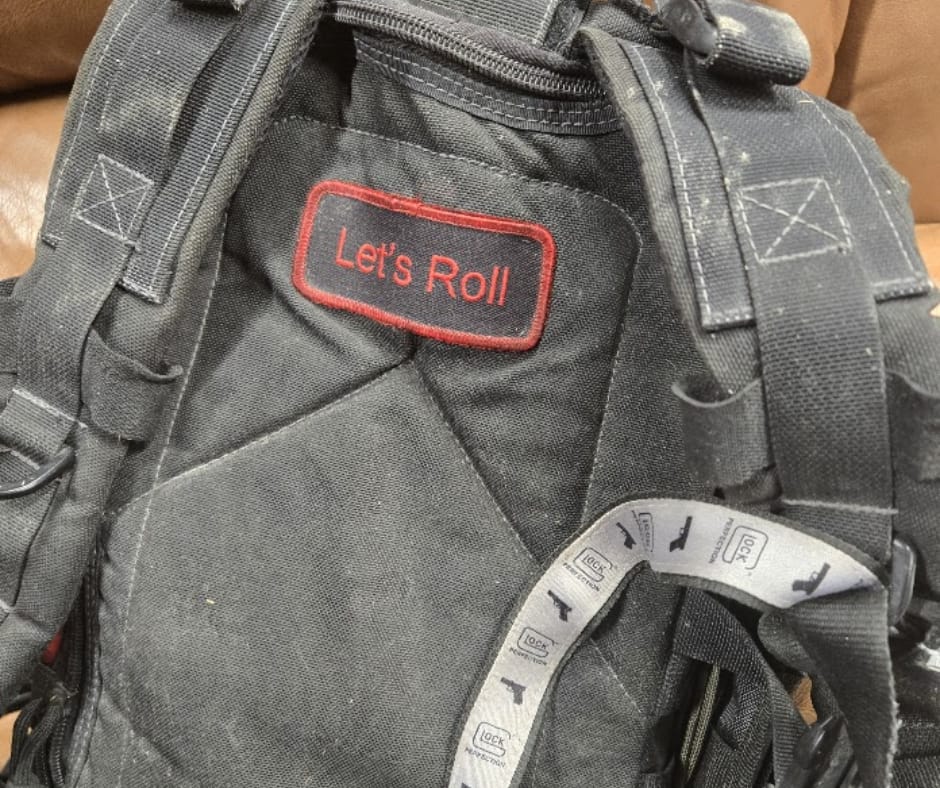 A close-up of a black tactical backpack featuring a red-bordered patch with the words "Let's Roll" in red text. A white ribbon with Glock logos is visible in the foreground, honoring the heroism of Todd Beamer and the passengers of United Flight 93.