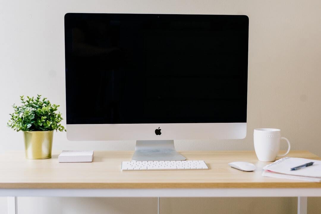 A clean white desk with an iMac displaying a black screen, white coffee mug, small potted plant, and minimal office supplies arranged neatly on a light wood surface