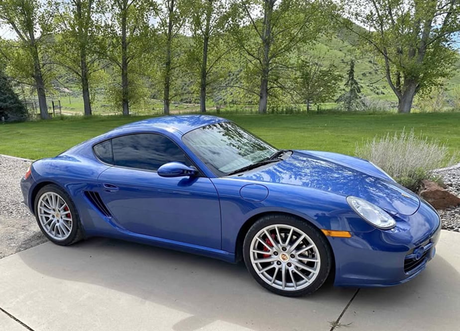 Side view of a cobalt blue 2007 Porsche Cayman S with silver wheels, parked in front of green hills