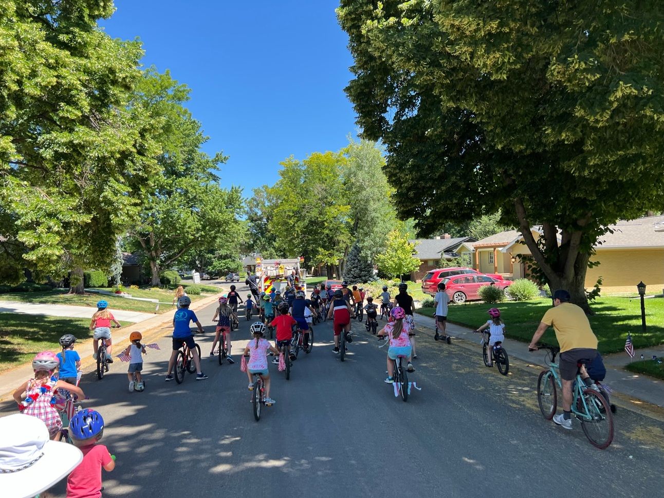 A photo of the backs of many kids and some adults on bikes following a fire truck down a tree-lined street.