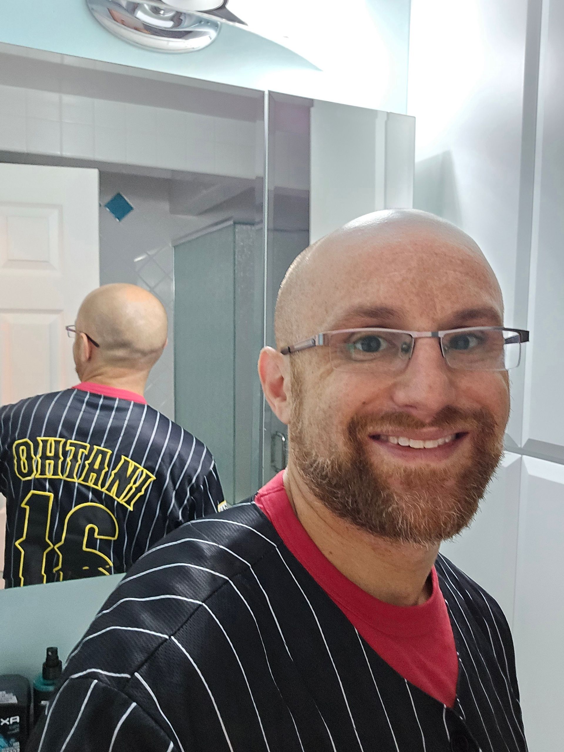 A selfie of me wearing an Ohtani jersey with black and white stripes over a red shirt, see in my bathroom mirror. I'm a bald, middle-aged, white man with a red beard flecked with white. I'm wearing glasses with a metal top rim.
