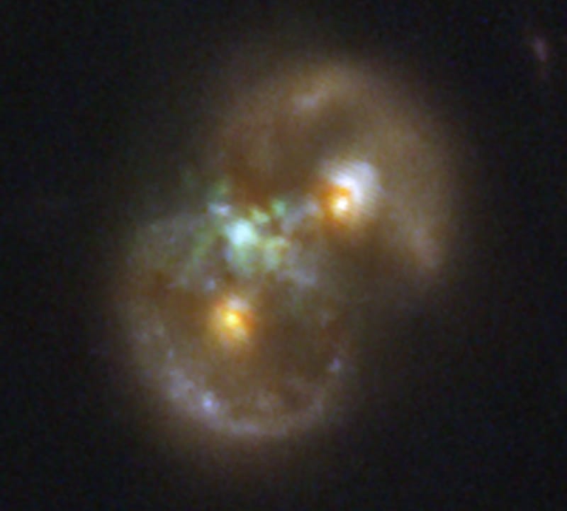 Two galaxies overlapping, looking a lot like two fried eggs next to each other in a pan. Two yellowish central blobs are surrounded by fuzzy rings, and offset from the center is a greenish blob of material.