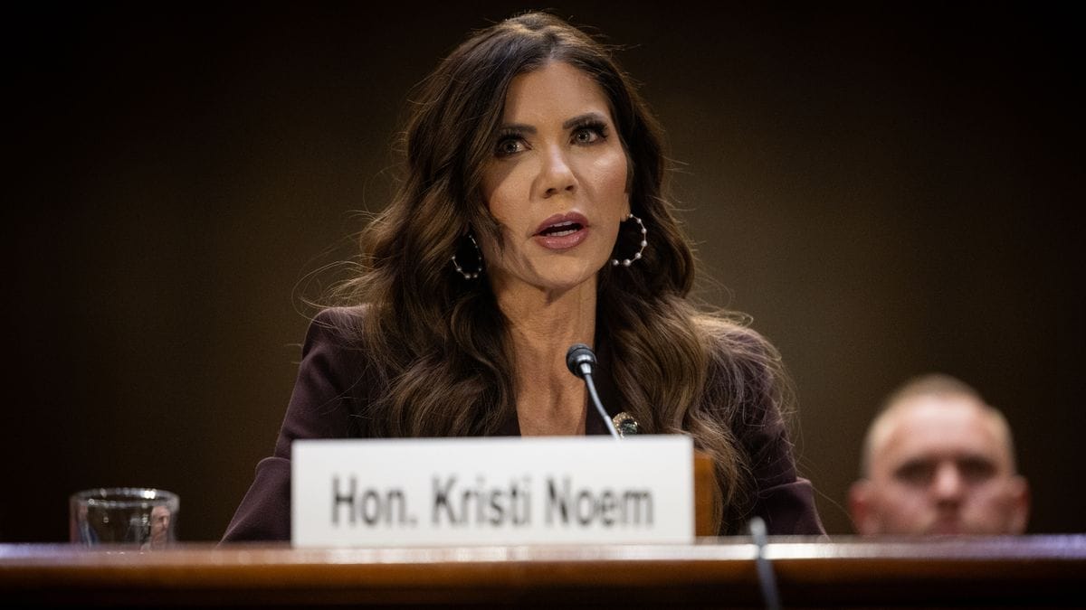 A white woman speaks while sitting at a desk. A plaque sitting in front of her says Hon. Kristi Noem. To her right, a person can be seen faintly sitting behind her.