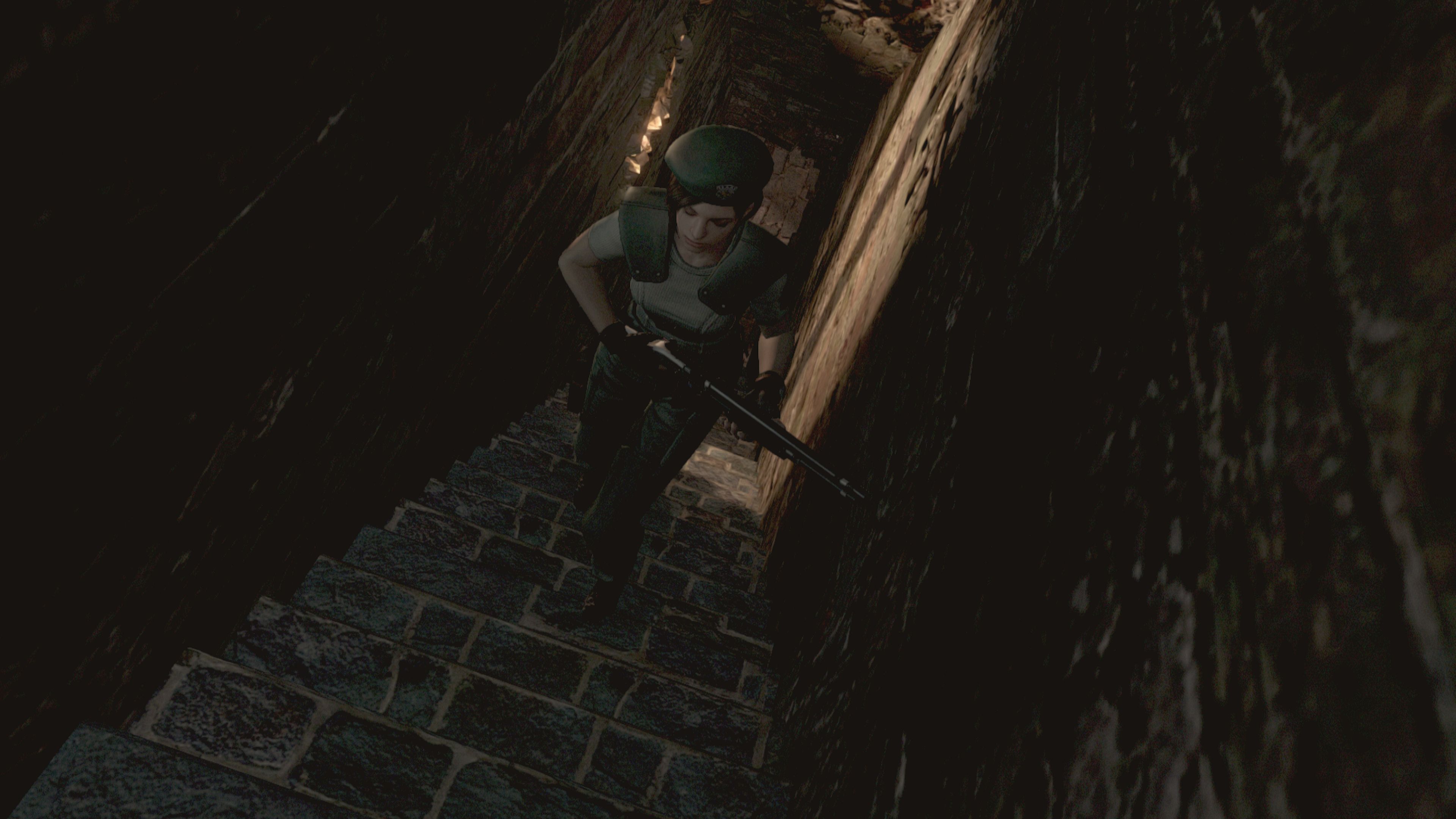 A screenshot from Resident Evil's 2002 remake, in its later HD edition, showing Jill Valentine with a shotgun in hand climbing up a long stairway. The fixed camera angle has her coming toward the player, up the stairs, and the anticipation of the climb (or descent) helps to build the tension of the moment that is only possible due to the lack of player control of what's seen.