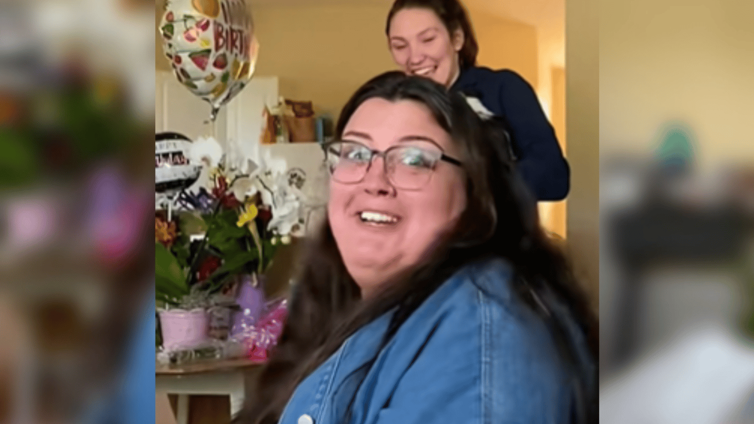A woman sits on a couch, looking over at the camera with a happily shocked expression on her face. A woman stands behind her, laughing. She's holding something that can't be seen in this image. A table to the side is full of plants and birthday balloons.