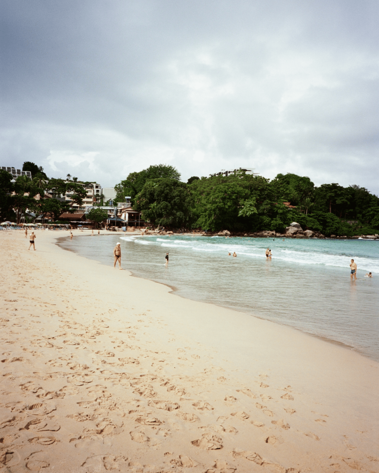 Kata beach with tourists enjoying the ocean with a resort and forested outcroping in the background.