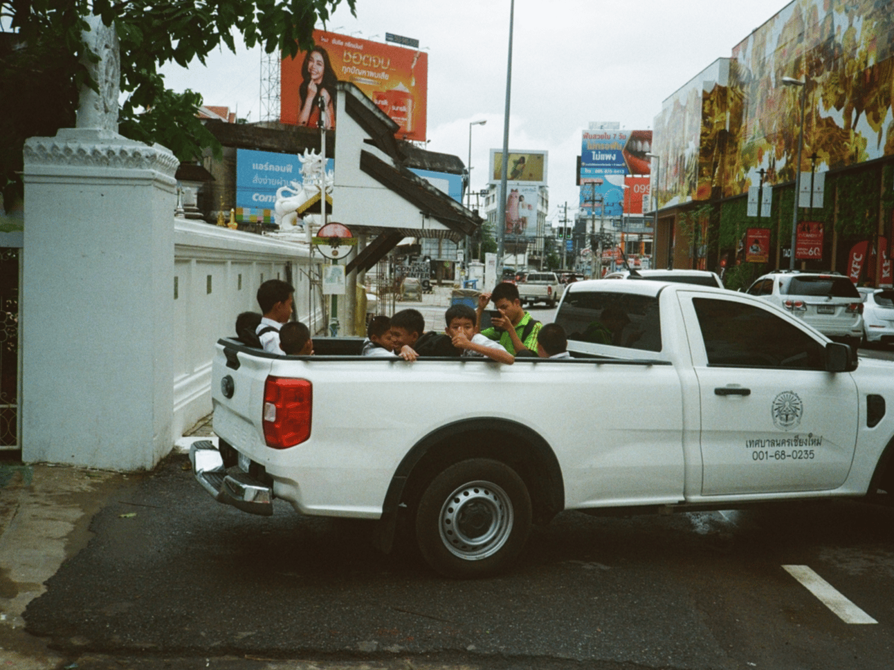 A group of school boys ride in the back of a white pickup. One looks at the camera and gives a thumbs up.