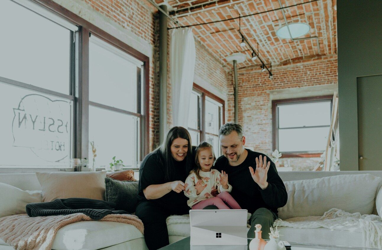 Parents sit on a white couuch at home with their daughter with an iPad in front of them, waving