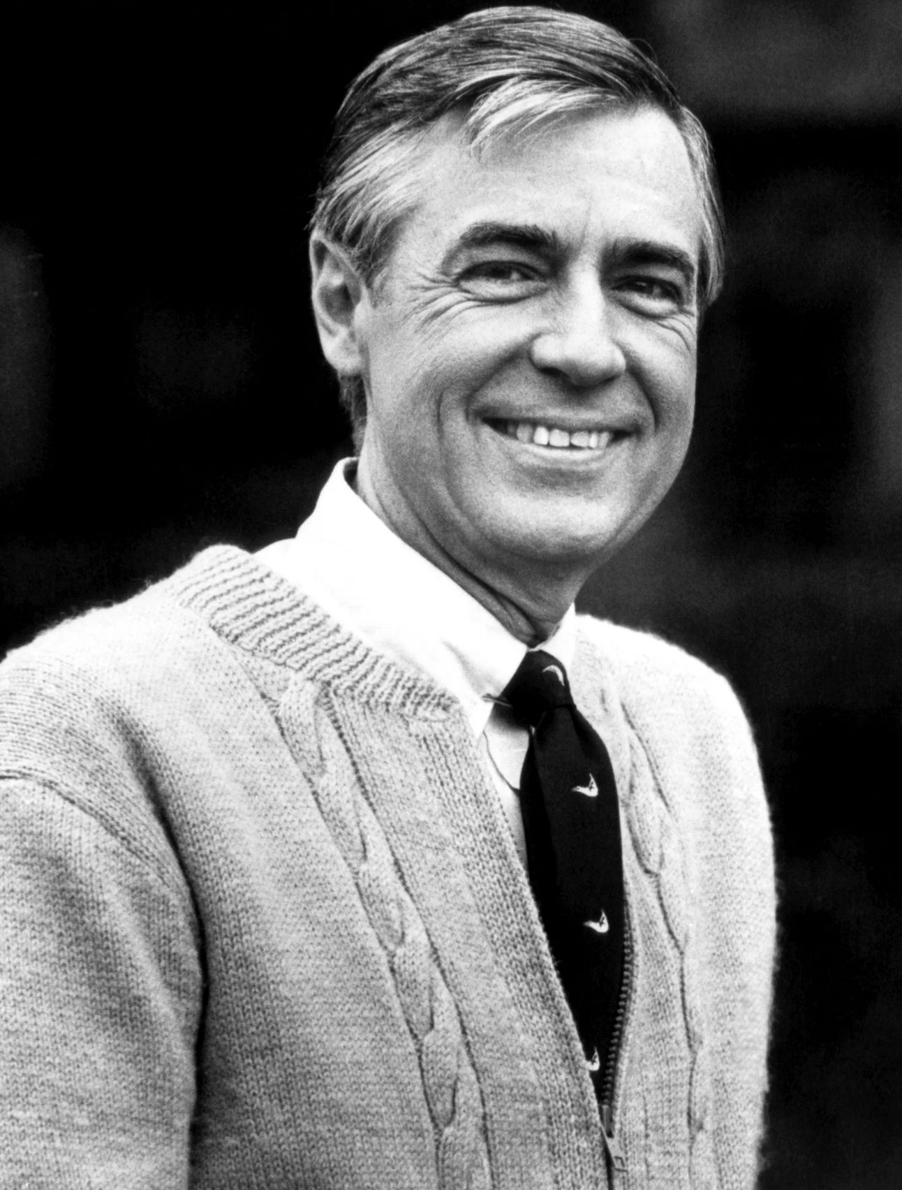 "Black and white portrait of Fred Rogers, smiling warmly. He is wearing a light cable-knit cardigan over a white shirt and dark tie, with a gentle, kind expression."