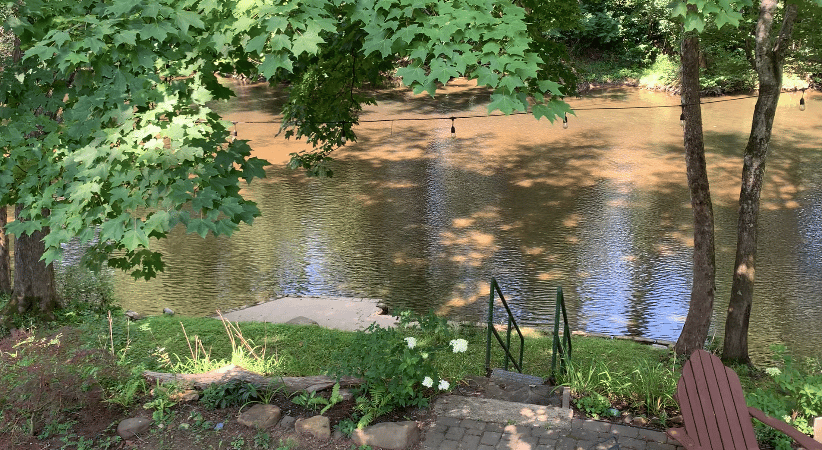 an idyllic landscape with part of a patio visible, and steps leading down to a small dock. Past it, the surface of a slightly muddy river is filled with tiny waves of motion. Higher up, trees in leaf are visible..